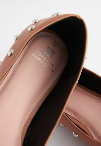 Brązowy but z ekoskóry z srebrnymi ćwiekami i wyściełanym różowym wnętrzem w rozmiarze 40, oznaczony "Forever Comfort."