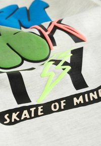 T-shirt en coton gris clair avec impression graphique multicolore : texte bleu et vert avec des accents néon rose et vert, et lettrage noir.