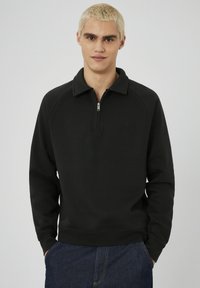 Jeune personne avec des cheveux blonds courts portant un sweat-shirt noir à zip quart et un jean bleu foncé, debout avec les mains dans les poches.