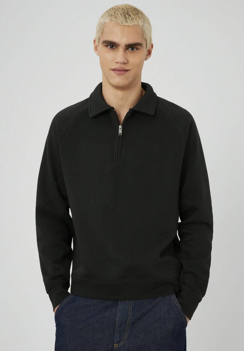 Jeune personne avec des cheveux blonds courts portant un sweat-shirt noir à zip quart et un jean bleu foncé, debout avec les mains dans les poches.