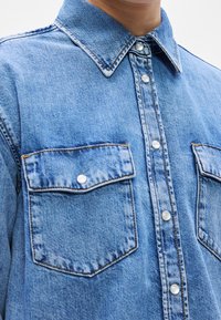 Chemise en denim bleu avec boutons pression en nacre, col pointu et deux poches avant à rabat avec fermeture par bouton.