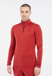 Crvena dugih rukava sportska majica s visokim ovratnikom i četvrtim patentnim zatvaračem. Sadrži suptilan logotip na prsima i elegantnu, glatku teksturu tkanine.