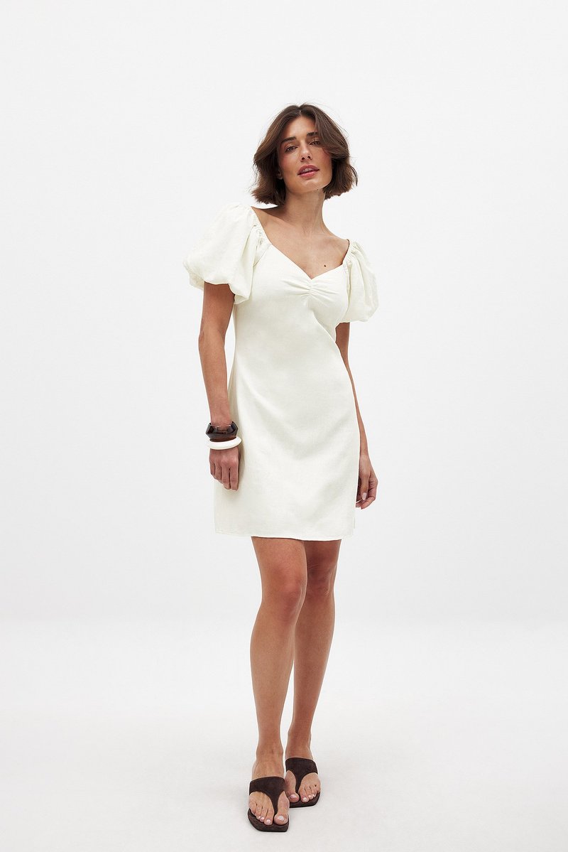 Weißes Leinen-Minikleid mit Puffärmeln, einer taillierten Körbchen und einer figurbetonten Silhouette. Dazu kombiniert mit braunen Sandalen und stapelbaren Armbändern.