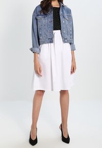 Giacca di jeans con un design cropped, colore blu; top nero a pois; gonna bianca a trapezio; décolleté nere con tacco a spillo. Texture morbide, stile casual.