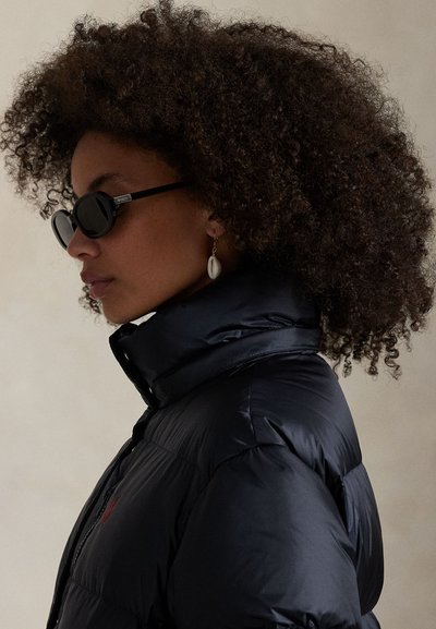 Profiel van een persoon met krullend haar die zwarte ovale zonnebrillen, een zwarte donsjacket en een schelpenoorbel draagt tegen een effen achtergrond.