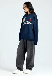 Femme debout portant un jean gris foncé large, des baskets blanches et un sweat-shirt bleu marine avec un cupidon, un cœur rouge et le texte "Je T'aime".