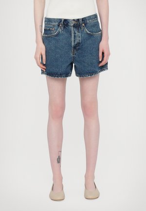 Shorts di denim blu scuro con una vestibilità classica, orlo grezzo, cinque tasche e metalleria in rame. Indossati con una maglietta bianca aderente e scarpe beige.