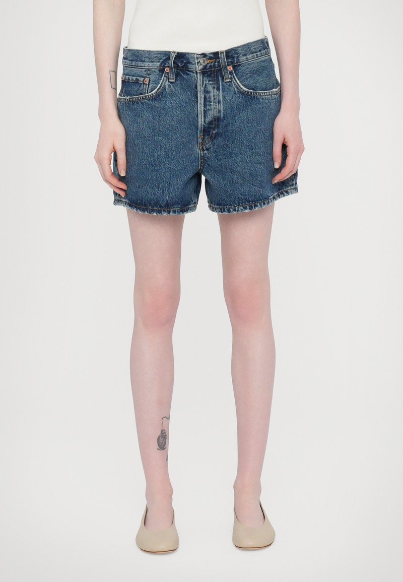 Shorts en denim bleu foncé avec une coupe classique, taille brute, cinq poches et accessoires en cuivre. Portés avec un haut blanc ajusté et des chaussures beiges.