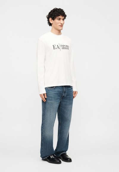 Νέος άντρας όρθιος, φοράει άσπρο πουκάμισο με μακριά μανίκια με το κείμενο "EA Emporio Armani", μπλε τζιν και μαύρα παπούτσια, σε απλό λευκό φόντο.
