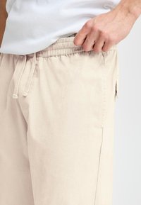 Beige shorts made of lightweight fabric, featuring an elastic waistband with drawstrings and side pockets. Smooth texture with no visible patterns.