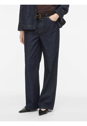Person iført mørkeblå vidde jeans med brun bælte, mørk jakke og sorte spidse sko, mod en ensfarvet lys baggrund.