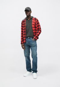 Camisa de flanela em xadrez vermelho e preto sobre um polo cinza, combinada com jeans azuis e ténis brancos, acessorizada com um boné preto.