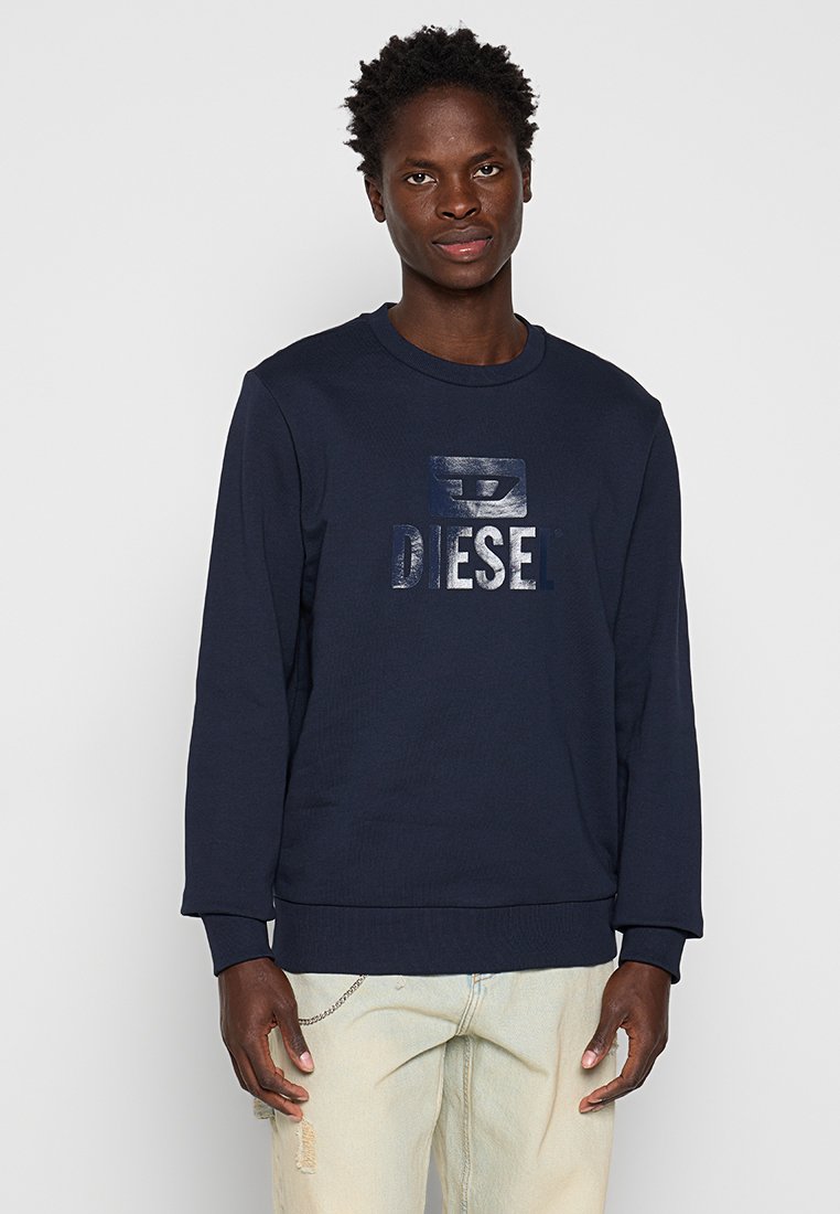 Giovane uomo che indossa una felpa blu navy con la scritta brillante "DIESEL" e jeans chiari beige strappati, in piedi contro uno sfondo chiaro uniforme.