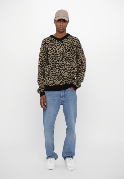 Leopard-print sweater with a black V-neckline, paired with relaxed blue jeans and white sneakers. The ensemble features a beige cap.