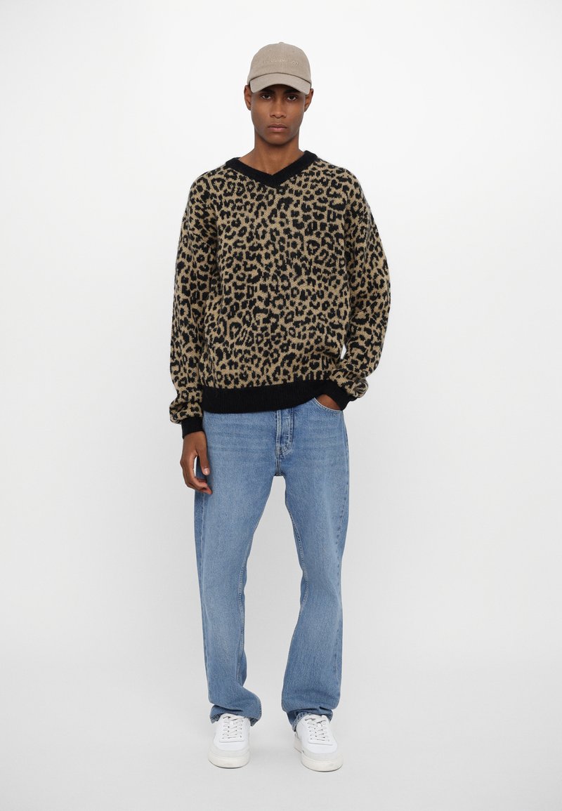 Leopard-print sweater with a black V-neckline, paired with relaxed blue jeans and white sneakers. The ensemble features a beige cap.