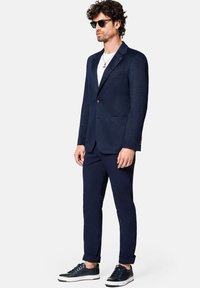 Granatowa marynarka o textured wykończeniu, z jednym guzikiem i dwiema przednimi kieszeniami, zestawiona z granatowymi spodniami i casualowymi trampkami.
