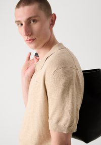 Polo din tricot bej cu mâneci scurte, având un finisaj texturat. O geantă neagră cu o suprafață netedă se află pe umăr.