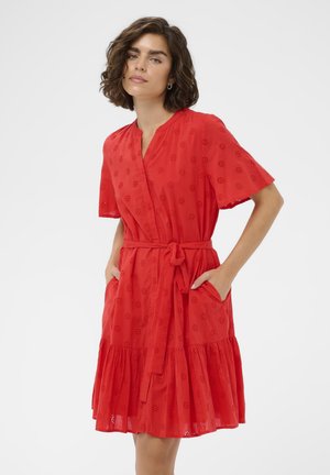 Mladá žena s krátkymi kučeravými vlasmi, oblečená v červenom krátkom šate s očkovými vzormi a vreckami, stojaca s rukami vreckách.