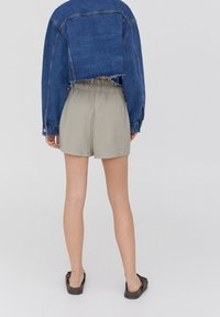 Veste en jean au design court, de couleur bleue, ourlet effiloché, associée à un short beige clair avec une ceinture élastique. Sandales marron visibles.