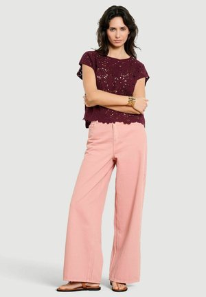 Donna in piedi con le braccia incrociate, indossa un top in crochet color amaranto, pantaloni larghi rosa, un braccialetto d'oro e sandali su uno sfondo semplice.