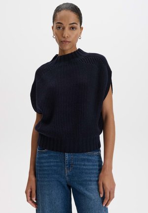 Vrouw draagt een zwarte gebreide mouwloze trui met geribde zoom en hoge hals, gecombineerd met blauwe denim jeans, staande tegen een effen achtergrond.