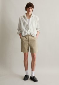 Striped button-up shirt in light beige and white, paired with khaki shorts. Model wears black loafers and white ankle socks, standing neutrally.