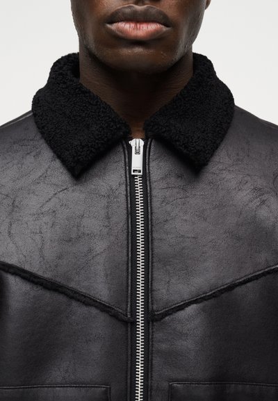Κοντινό πλάνο ενός ατόμου που φοράει μαύρο δερμάτινο μπουφάν με μαύρο κολάρο από φλις και ασημένιο μπροστινό φερμουάρ.