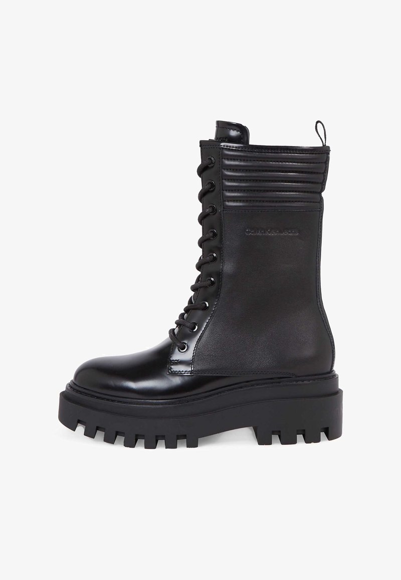 Černá kotníková bota na šněrování s lesklým koženým povrchem, žebrovaným detailem na vrchu a robustní podrážkou s výrazným vzorkem.