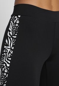 Svarta leggings med en sidopanel som har ett kontrasterande abstrakt vitt mönster. Materialet verkar slätt, stretchigt och åtsittande.