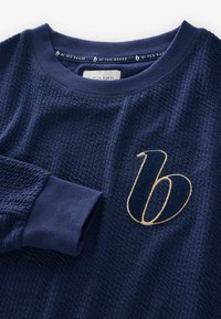 Marineblauwe sweatshirt van textuurstof, met geribbelde manchetten en een gouden geborduurd accent van de letter 'b' op de voorkant. Donkere contrasterende halslijn.