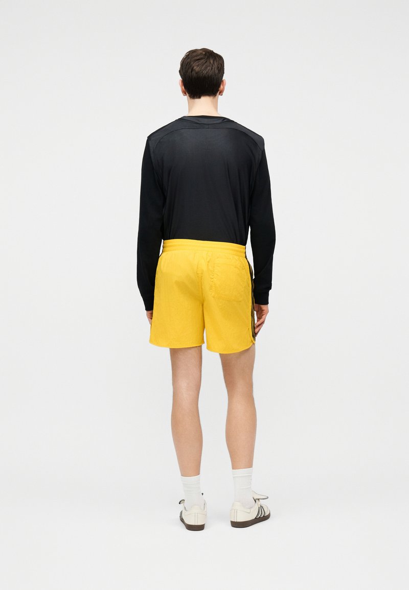 Mand stående med fronten væk fra kameraet iført sort langærmet skjorte, gule shorts med sidelængder, hvide sokker og hvide sneakers på ensfarvet baggrund.