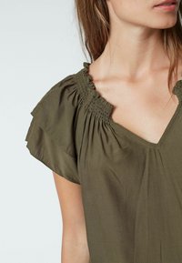 Donna che indossa una blusa verde oliva con scollatura arricciata ed elastica e maniche a volant, accorciata per mostrare l'area delle spalle e del collo.