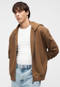 Καφέ φούτερ με φερμουάρ, υφασμένο με υφή, μεγάλο κουκούλα και δύο μπροστινές τσέπες, συνδυασμένο με λευκό t-shirt και ανοιχτό μπλε τζιν.