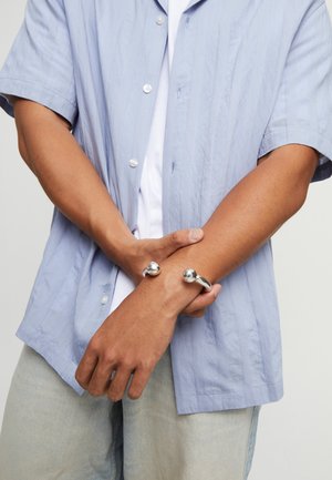Chemise bleue claire à manches courtes avec des boutons argentés, portée par-dessus un t-shirt blanc. Un bracelet métallique avec deux accents sphériques au poignet.