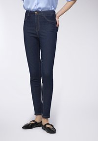 Jeans in denim blu scuro con vita alta, fit dritto, dettagli di cuciture visibili e tasche frontali. Abbinati a scarpe nere a pianta piatta.