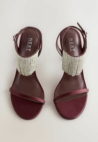 Burgunderfarbene Sandalen mit einem strukturierten Riemen, der mit silbernen Perlen verziert ist. Sie besitzen ein Slingback-Design und eine flache, glatte Sohle.