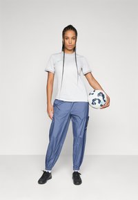 Une femme en t-shirt de sport blanc et pantalon bleu tient un ballon de football sur le côté, se tenant devant un fond clair et uni.