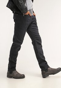 Jeans negros ajustados de corte slim, hechos de mezclilla, con una textura suave y detalles mínimos, combinados con botines grises. El modelo está en una pose caminando.