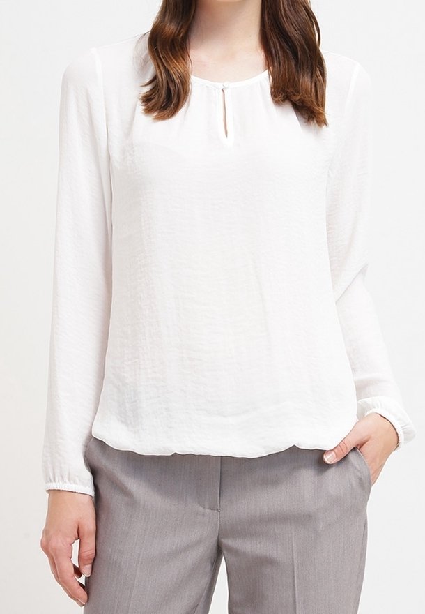 Femme portant une blouse blanche à manches longues avec un petit col fendu boutonné et un pantalon gris, une main dans la poche.