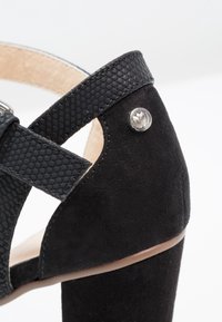 Sandale noire à talons hauts avec des sangles en cuir texturé, des accents en daim, un talon épais et une breloque en argent décorative à l'arrière.