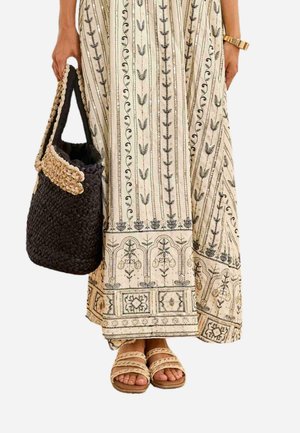 Mujer que lleva un vestido largo color crema con estampados florales negros, sosteniendo un bolso negro tejido y usando sandalias beige con tiras trenzadas.