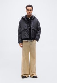 Νεαρός άνδρας που φοράει μαύρο μπουφάν puffer, μαύρο πουκάμισο, μπεζ παντελόνι με φαρδιά πόδια και μαύρα παπούτσια, στέκεται μπροστά σε ένα απλό λευκό φόντο.