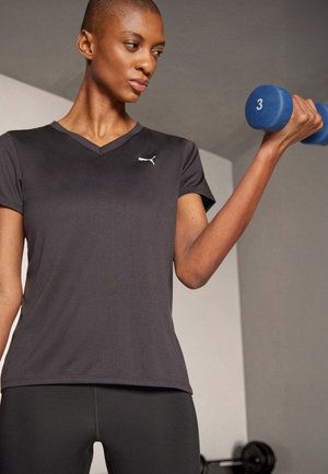 Tricou sportiv negru cu decolteu în V și mâneci scurte, având un mic logo alb. Modelul ține o gantera albastră. Design slim-fit.