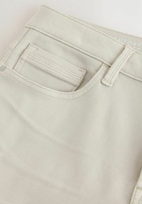 Nahaufnahme der vorderen Tasche einer beigen Hose mit Gürtelschlaufe und sichtbaren Nahtdetails.