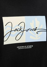 Sort tekstil T-shirt med et rektangulært grafik, der har lyseblå og cremefarvede elementer, sort kursiv tekst "Jack & Jones" og hvid tekst nedenfor.
