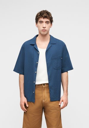 Tânăr în picioare, purtând o cămașă albastră cu mânecă scurtă pe nasturi peste un maiou alb și pantaloni scurți maro.