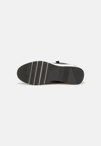 Semelle d'une basket noire et grise avec des motifs de grip texturés et le nom de la marque "Tamaris" embossé près de la zone de la voûte plantaire.