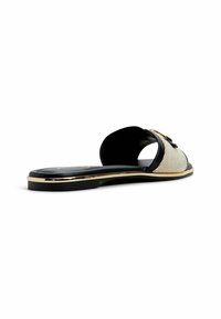 Slide sandal with a woven cream upper and black accents, featuring a gold-tone band along the sole and a decorative embellishment on the upper.