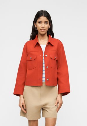 Femme aux longs cheveux noirs portant des boucles d'oreilles créoles, une veste rouge boutonnée sur une chemise rayée et un short beige, debout devant un fond blanc uni.