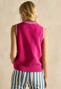 Rozā adīta bezpiedurkņu blūze ar tekstūru un baltu ribotu apkakli, saskaņota ar svītrainām džinsu biksēm. Attēlota aizmugures skats.